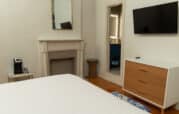 Minimalist bedroom with a bed, TV on the wall, dresser, coffee machine, small mirror, and a non-functional fireplace. An open door leads to a bathroom with a partially visible sink.