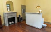 A room with yellow walls features a fireplace, a small black shelf, and a white reception desk with a gold lamp and tissue box on top. The floor is wooden.