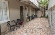 Brick courtyard with outdoor seating, featuring black metal tables and chairs. Potted plants are placed along the walls, and palm trees are visible in the background.