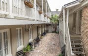 A narrow courtyard with brick flooring is flanked by beige buildings. Black chairs and tables, along with potted plants, line the sides. Wrought-iron baskets hang above.