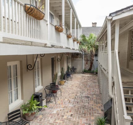 A narrow courtyard with brick flooring is flanked by beige buildings. Black chairs and tables, along with potted plants, line the sides. Wrought-iron baskets hang above.