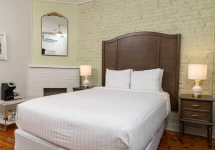 A neatly made bed with white linens, flanked by two wooden nightstands with lamps, positioned against a light-colored brick wall. A mirror and a non-functional fireplace are also visible.