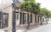 A beige building with dark shutters and a lantern stands on a brick sidewalk. Trees line the street in front of it.