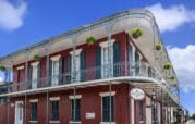 A red two-story building with ornate wrought iron balconies and hanging plants on the corner of a street under a blue sky.