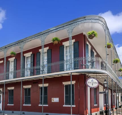 A red two-story building with ornate wrought iron balconies and hanging plants on the corner of a street under a blue sky.