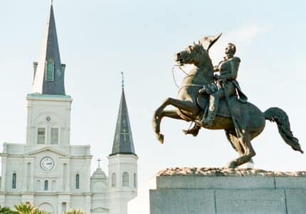 Equestrian statue of a man in front of a historic cathedral with two towers and a clock on one. The sky is clear.