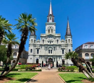 Saint Louis Cathedral in New Orleans, with its three spires, is surrounded by palm trees and a green lawn under a clear blue sky.