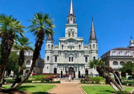 Saint Louis Cathedral in New Orleans, with its three spires, is surrounded by palm trees and a green lawn under a clear blue sky.