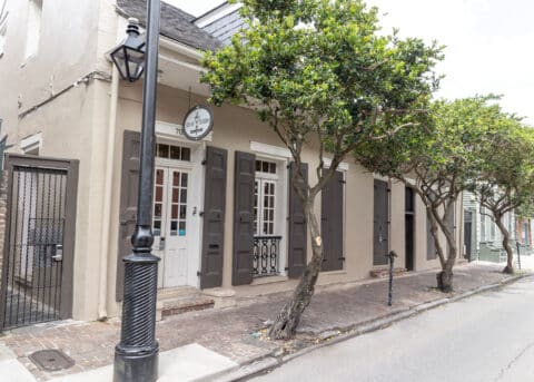 Historic building with beige stucco walls, dark shutters, and a sign on a quiet street lined with trees, a black lamppost, and a gated entrance.