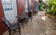 A narrow brick patio with black wrought iron tables and chairs against a red building, lined with palm plants and greenery.
