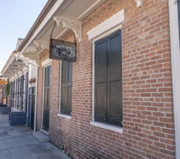 Sidewalk view of a brick building with a sign for apartments. Black shutters cover tall windows. Cars are parked along the street, and ornate brackets decorate the eaves.