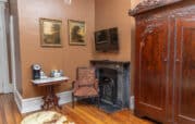 Room with a marble-topped table, coffee machine, chair, fireplace, two paintings, wooden wardrobe, and wall-mounted TV. Wood flooring and brown walls.