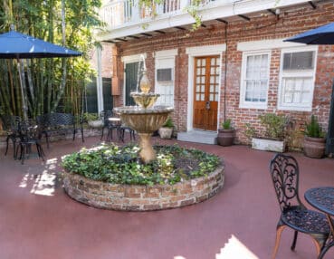 A courtyard featuring a central stone fountain, surrounded by potted plants and wrought iron patio furniture, with a brick building in the background.