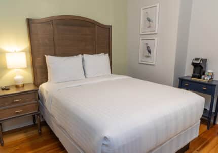 A neatly made bed with white linens is flanked by wooden nightstands. A lit lamp and coffee maker sit on the tables. Two bird illustrations hang on the pale green wall.