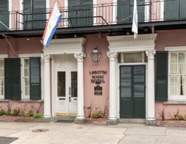 Facade of the Lafitte House Hotel featuring pink walls, green shutters, three flags, and white columns on a balcony.