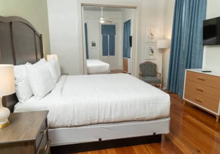 Hotel room with a large bed, white linens, two bedside tables with lamps, a dresser with a TV, a chair, and blue curtains. Reflective mirror on the back wall.