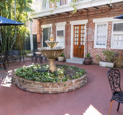 Brick courtyard with a central stone fountain, surrounded by potted plants and wrought iron chairs and tables. Two large umbrellas provide shade. Red brick building in the background.