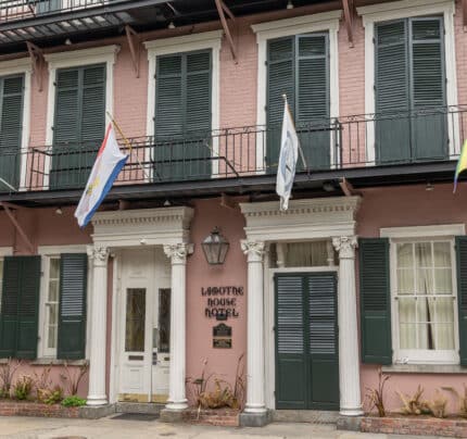 Three-story pink building with green shutters, named "Lafitte House Hotel." Flags hang above the entrance.