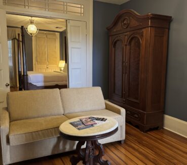 A room with a beige sofa, round wooden table with magazines, large wooden wardrobe, chandelier, wooden floors, and a white door leading to a bed with a lit lamp in the background.