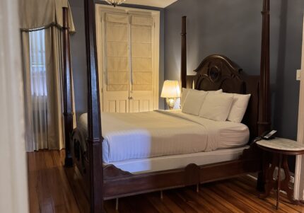 Bedroom with a four-poster wooden bed, white bedding, and a lit lamp on a bedside table. Dark blue walls, wooden floor, chandelier, and sheer drapes on a window.