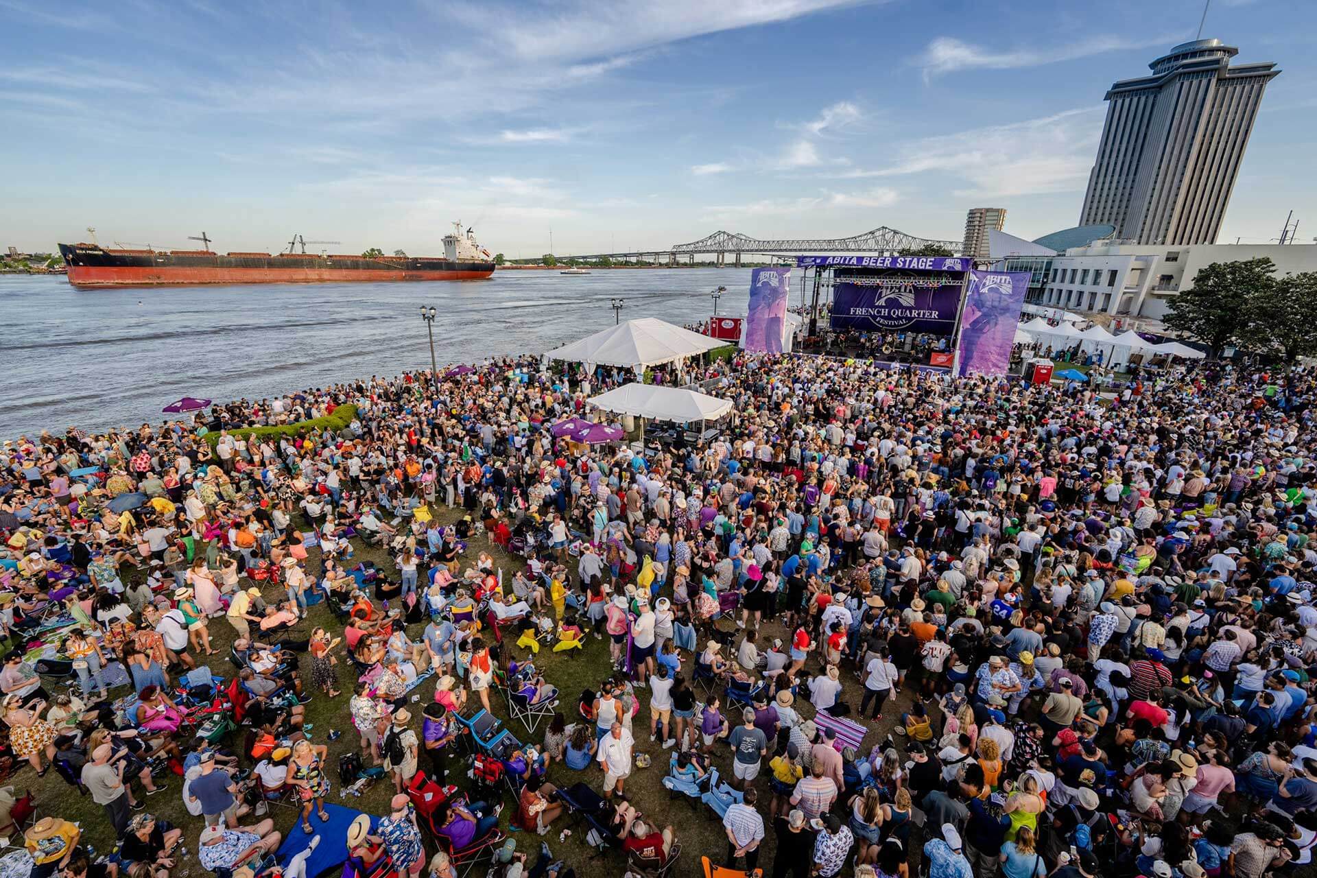 Lively outdoor festival by the river with crowds, a large stage, and a scenic bridge view, ideal for event-loving hotel guests.
